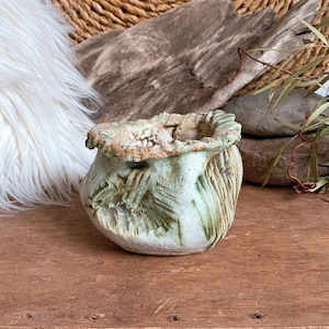 May include: A small, handcrafted ceramic pot with a textured, organic design. The pot features a light green and brown color palette, with visible brushstrokes and a rough, uneven rim. The exterior has a carved leaf pattern. The pot is displayed on a wooden surface.