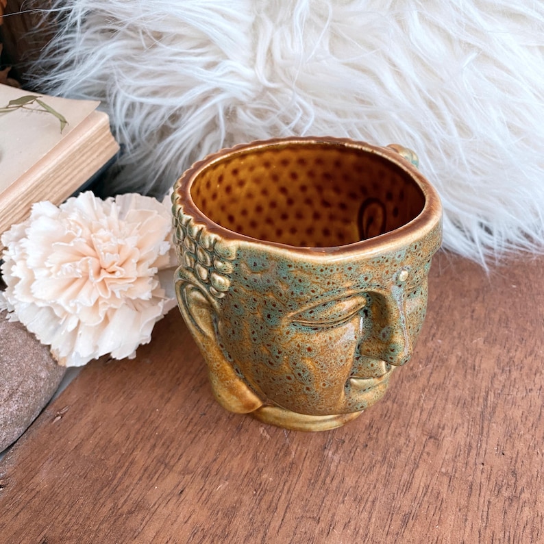 May include: A ceramic Buddha head planter with a textured, speckled interior and a detailed exterior. The planter features a blend of brown, green, and gold tones, with the Buddha's face in a serene expression. The planter is designed to hold small plants or succulents.