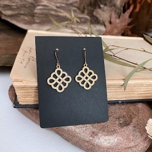 Peut inclure: Une paire de boucles d'oreilles dorées avec un motif de nœud décoratif. Chaque boucle d'oreille présente une série de boucles imbriquées, incrustées de petites pierres scintillantes. Les boucles d'oreilles sont présentées sur une carte noire, sur un fond de livre ancien et d'éléments naturels.