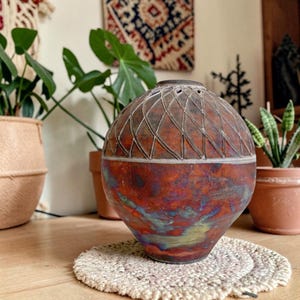 May include: A spherical ceramic vase featuring a dark, iridescent finish with swirling hues of copper, blue, and green. The upper portion displays a carved, repeating diamond lattice pattern, while the lower section remains smooth and polished.