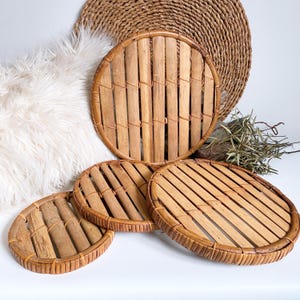 May include: A set of four round, natural bamboo coasters. The coasters have a woven design with vertical bamboo slats held together by a woven border. A larger, similar woven mat is in the background. The coasters are a light brown color.