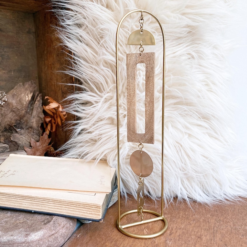 May include: A gold-toned metal arch stand featuring a vertical hanging mobile. The mobile consists of a brass semi-circle, a rectangular wooden frame, a circular brass disc, and a small brass rectangular piece, all connected by thin metal rings.