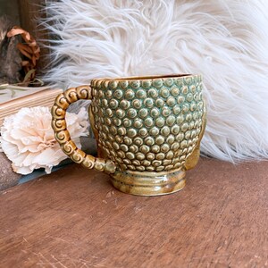 May include: A ceramic mug with a textured, dotted surface in shades of green and brown. The handle is a detailed, looped design. The mug is set on a wooden surface, with a fluffy white background.