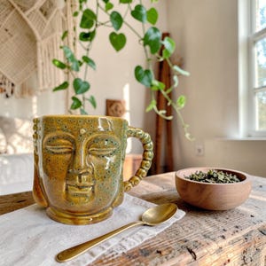 May include: A ceramic mug shaped like a Buddha head with a glossy mustard yellow and green speckled glaze. The mug features a textured handle, a small wooden bowl filled with loose tea leaves, and a gold-toned metal spoon resting on a white cloth.