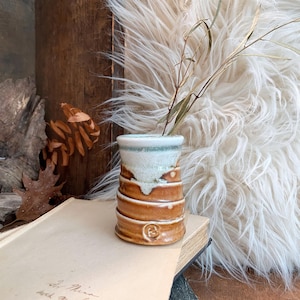 May include: A small, cylindrical ceramic vase with a glossy finish, featuring a warm brown base that transitions into a creamy white glaze at the top. The exterior has horizontal ridges and a small stamped circular mark near the bottom edge.