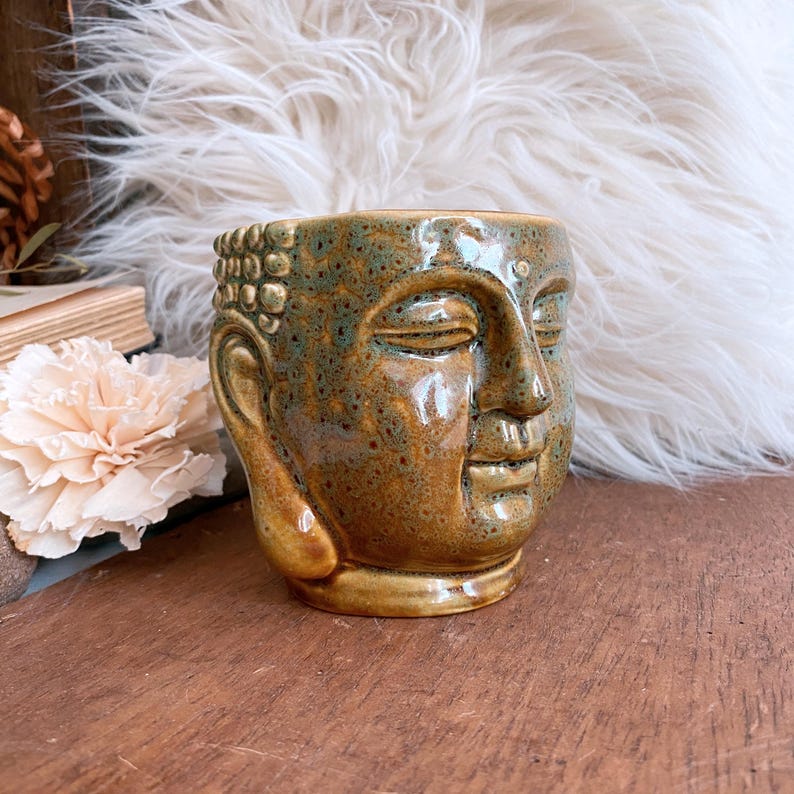 May include: A ceramic Buddha head planter in shades of brown and green. The planter has a detailed face with closed eyes and a serene expression. The surface has a speckled texture, and the overall design is reminiscent of a traditional Buddhist sculpture.