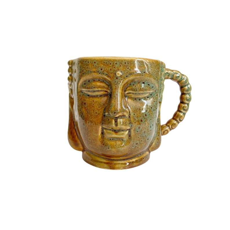 May include: A ceramic mug shaped like a Buddha head with a glossy, speckled mustard-yellow and green glaze. The mug features closed eyes, a serene facial expression, and a handle textured with a series of small, rounded, bead-like ceramic shapes.