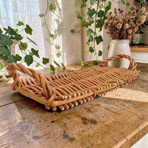 May include: A rectangular woven wicker tray featuring a light brown natural finish, a flat base, and two braided handles on the ends. The tray is constructed from thin, flexible willow branches arranged in a parallel pattern with a sturdy border.