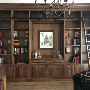 May include: A dark wood bookcase with a ladder, a framed picture, and a small bar area. The bookcase has multiple shelves filled with books.