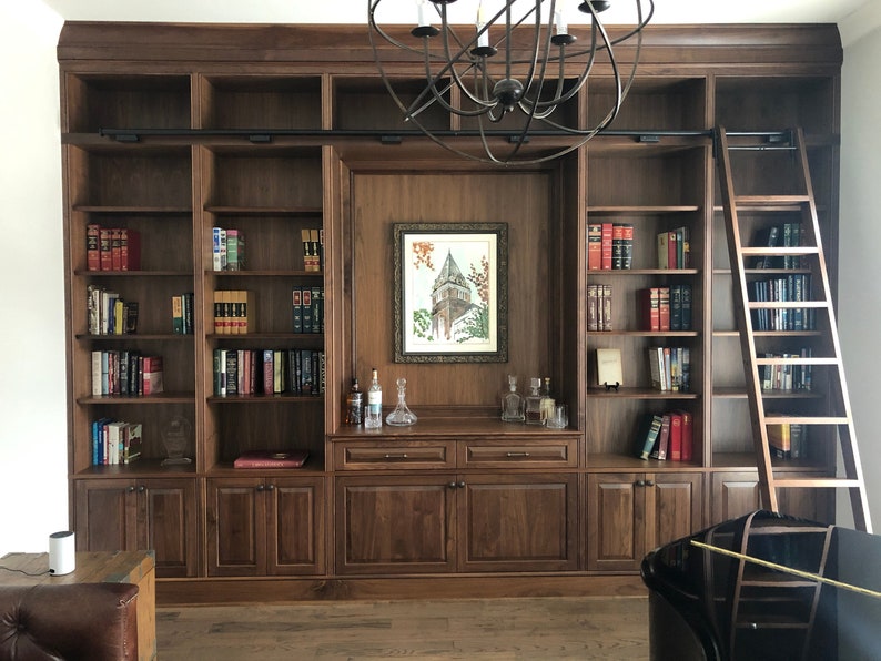 May include: A dark wood bookcase with a ladder, a framed picture, and a small bar area. The bookcase has multiple shelves filled with books.