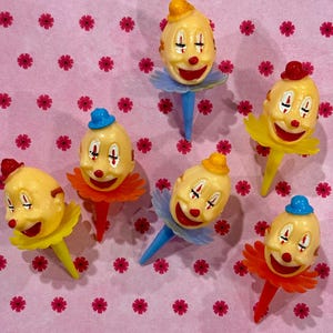 May include: A collection of vintage clown cake toppers. Each topper features a smiling clown face with painted details, a colorful ruffled collar, and a pointed base. The clowns wear small hats in various colors.