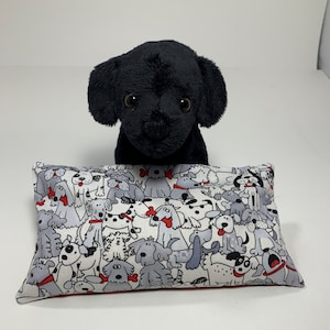 May include: A black plush dog toy sitting on a white pillow with a pattern of black and white dogs. The pillow has a red border.
