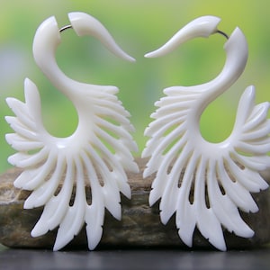 Puede incluir: Un par de pendientes de hueso blanco con un diseño detallado e intrincado que se asemeja a plumas. Los pendientes tienen forma de gancho y presentan un patrón delicado y calado.