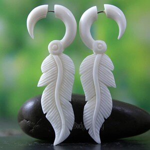 Puede incluir: Un par de pendientes de plumas de hueso blanco. Los pendientes tienen forma de pluma con una pieza de hueso curvada en la parte superior. Los pendientes cuelgan de una piedra negra.