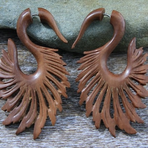 Puede incluir: Un par de pendientes de madera marrón con un diseño tallado que se asemeja a plumas. Los pendientes tienen forma de gancho curvo con un diseño de ala decorativo.