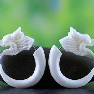 May include: A pair of white bone dragon head earrings. The earrings are shaped like a dragon's head with a curved bone hoop. The earrings are made of bone and are a tribal style.