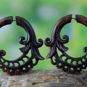 Puede incluir: Un par de pendientes de madera marrón con un diseño floral tallado. Los pendientes tienen forma de media luna y presentan un patrón delicado e intrincado.