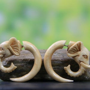 Puede incluir: Un par de pendientes con forma de elefante de color marfil. Cada pendiente presenta una cabeza de elefante detallada y un diseño curvo en forma de colmillo. Los pendientes se exhiben sobre una piedra contra un fondo verde borroso.