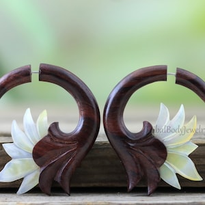 May include: A pair of dark brown wooden earrings with a curved design, featuring a carved floral accent in white and pale yellow. The earrings have a post closure. Set against a blurred green background.