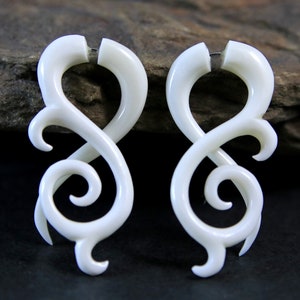Puede incluir: Par de pendientes de hueso blanco con un diseño intrincado y en espiral. Los pendientes tienen forma de ocho y un acabado liso y pulido.