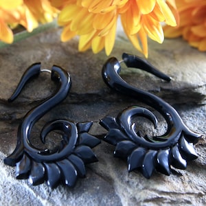May include: A pair of black, carved, wooden ear gauges with a floral design. The gauges are shaped like a curved horn with a floral design at the end.