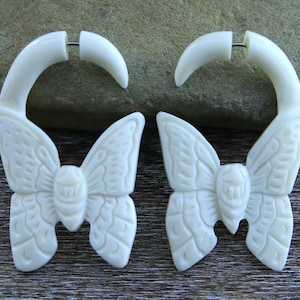 May include: A pair of white bone butterfly earrings with a carved design. The earrings are shaped like a butterfly with a curved hoop that goes through the ear.
