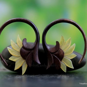 May include: A pair of wooden ear gauges with a carved sunflower design. The gauges are brown and the sunflowers are yellow.