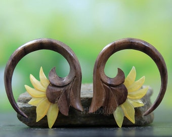 May include: A pair of wooden ear gauges with a sunflower design. The gauges are made of dark wood and have a yellow and white sunflower carved into the side. The gauges are shown on a rock.