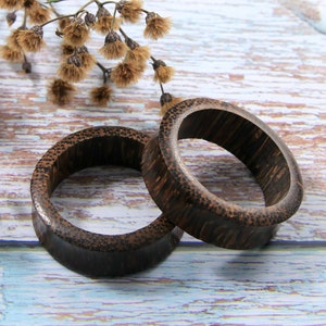 May include: Two dark brown, wooden ear gauges with a textured surface. The gauges are oval-shaped and appear to be made of a dark wood. A sprig of dried flowers is in the background.