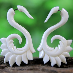 Puede incluir: Un par de pendientes de hueso blanco con forma de pájaro estilizado con un diseño floral. Los pendientes están tallados de forma intrincada y tienen un aspecto delicado y femenino.