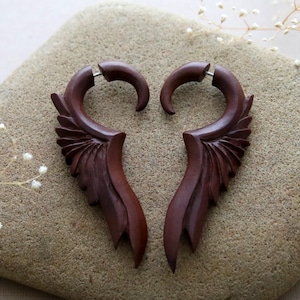 Puede incluir: Un par de pendientes de madera marrón oscuro. Cada pendiente presenta una parte superior curvada y un diseño en forma de ala con tallas detalladas de plumas. Los pendientes se muestran sobre una superficie texturizada de color marrón claro.