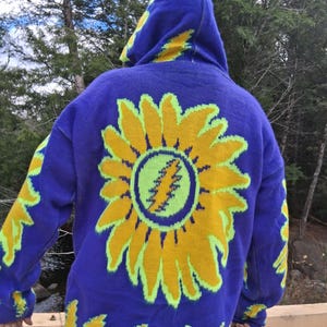 May include: A blue hooded sweater with a large yellow sunflower design on the back. The sunflower has a green lightning bolt in the center. Yellow accents are on the sleeves and hood. The sweater is made of a knitted material.