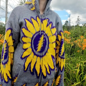 May include: Gray zip-up hoodie with a large yellow sunflower design and a purple lightning bolt graphic. The hoodie has a hood and pockets.