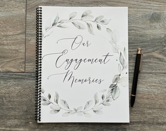 Puede incluir: Un cuaderno blanco de espiral con una corona de hojas verdes en acuarela y el texto "Our Engagement Memories" en cursiva negra.