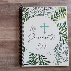 May include: A spiral-bound notebook with a white cover featuring the text "My Sacraments Book" in elegant script, a teal cross, and a watercolor design of green leaves. The notebook is on a wooden surface.