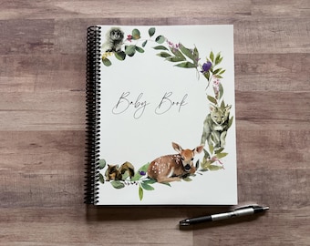 May include: A spiral-bound baby book with a white cover featuring a watercolor design. The cover includes the text "Baby Book" in script, surrounded by a wreath of greenery, an owl, a fawn, a coyote, and other forest animals.