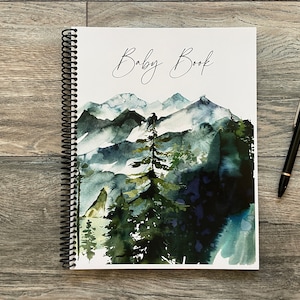 May include: A spiral bound baby book with a watercolor illustration of a mountain range with trees and a white background. The title "Baby Book" is written in cursive on the front cover.
