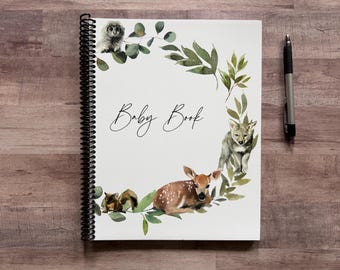 May include: A spiral-bound baby book with a white cover featuring watercolor illustrations of woodland animals, including an owl, deer, and fox, surrounded by green foliage. The words "Baby Book" are written in elegant script. A pen rests beside the book.