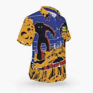 May include: A blue and yellow short-sleeved button-down shirt with a graphic print of a monster on a planet. The shirt has a pocket on the left side with the text "Space Wanderers" printed on it.
