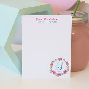 May include: A white notepad with the text "From the desk of: Mrs. Scragg" in pink and teal. The notepad features a floral wreath with the letter "S" in the center. A pink jar with a daisy and a geometric mint green shape are in the background.