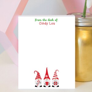 Puede incluir: Un bloc de notas blanco con el texto "From the desk of: Cindy Lou" en verde y rojo. La parte inferior del bloc de notas presenta tres gnomos navideños con sombreros rojos y sosteniendo regalos. Un tarro dorado está en el fondo.
