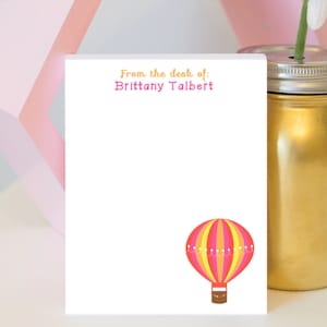 May include: White notepad with the text "From the desk of: Brittany Talbert" at the top. A pink and yellow hot air balloon with a brown basket is at the bottom of the notepad.