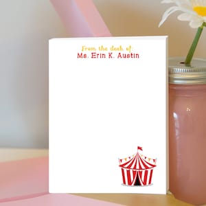 Puede incluir: Bloc de notas blanco con texto rojo que dice "From the desk of: Ms. Erin K. Austin". Una carpa de circo roja y blanca a rayas con una bandera amarilla se encuentra en la parte inferior del bloc de notas.