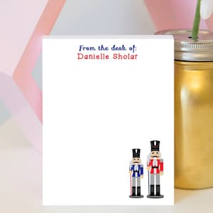 May include: White notepad with the text "From the desk of: Danielle Sholar" at the top. The bottom of the notepad features two cartoon nutcracker figures, one in blue and one in red.