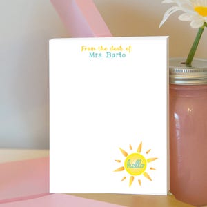 Puede incluir: Bloc de notas blanco con el texto "From the desk of: Mrs. Barto" en la parte superior. Un sol amarillo con un "hello" azul en su interior se encuentra en la parte inferior del bloc de notas.