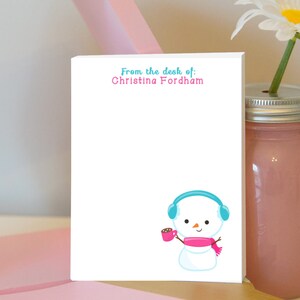May include: A white notepad with the text "From the desk of: Christina Fordham" at the top. A cartoon snowman with a blue headband and pink scarf is holding a pink mug with a brown drink inside.