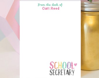 May include: A white notepad with the text "From the desk of: Cari Reed" in teal and pink. The bottom of the notepad has the words "SCHOOL SECRETARY" in colorful, dotted letters and a pink heart. A gold jar is in the background.