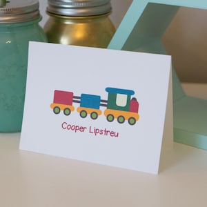 May include: A white card with a colorful cartoon train and the text "Cooper Lipstreu". The card is sitting on a white surface next to a green mason jar and a gold mason jar.
