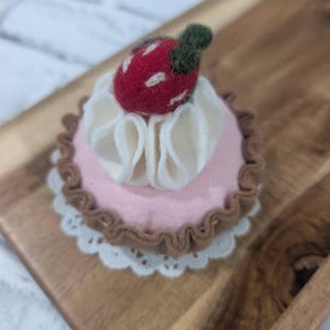 May include: A felt strawberry tart with a pink base, white cream topping, and a red strawberry with green leaves. The tart sits on a white lace doily and a brown wooden tray. A charming, handmade dessert.
