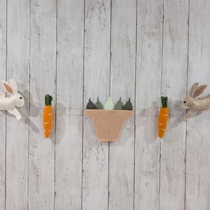 May include: A felt garland with two white bunnies, two orange carrots, and a green plant in a brown pot. The garland is strung on a white string.